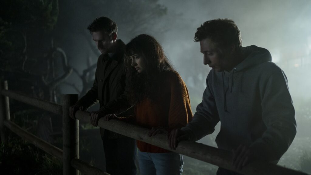 The Rental movie scene. Three people stand by a wooden fence at night, looking down with concerned expressions. Suspenseful thriller.