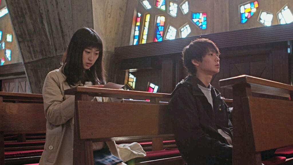 Sacrifice | Taku Tsuboi Sacrifice film still: Two young people sit in a church pew. Stained glass windows are visible.