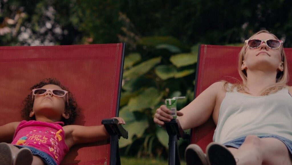 Saint Frances film still: Woman and child in sunglasses relax in lawn chairs, enjoying the sun. Juice box.