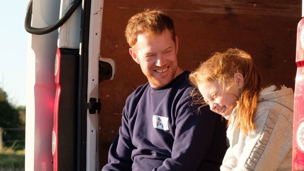 Sorry We Missed You film still. A delivery driver and his daughter smile in a van. Ken Loach film.