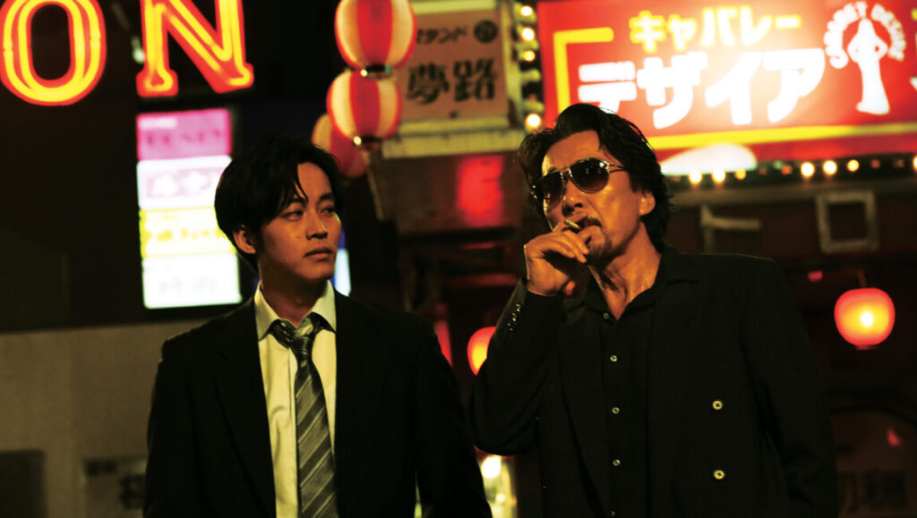 The Blood of Wolves film still. Two men in suits stand in a neon-lit street. One smokes a cigarette.