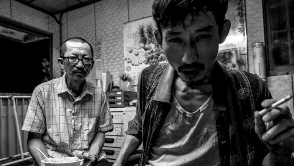 Black and white still from The Great Buddha+, featuring two men indoors, one smoking. Huang Hsin-yao film.