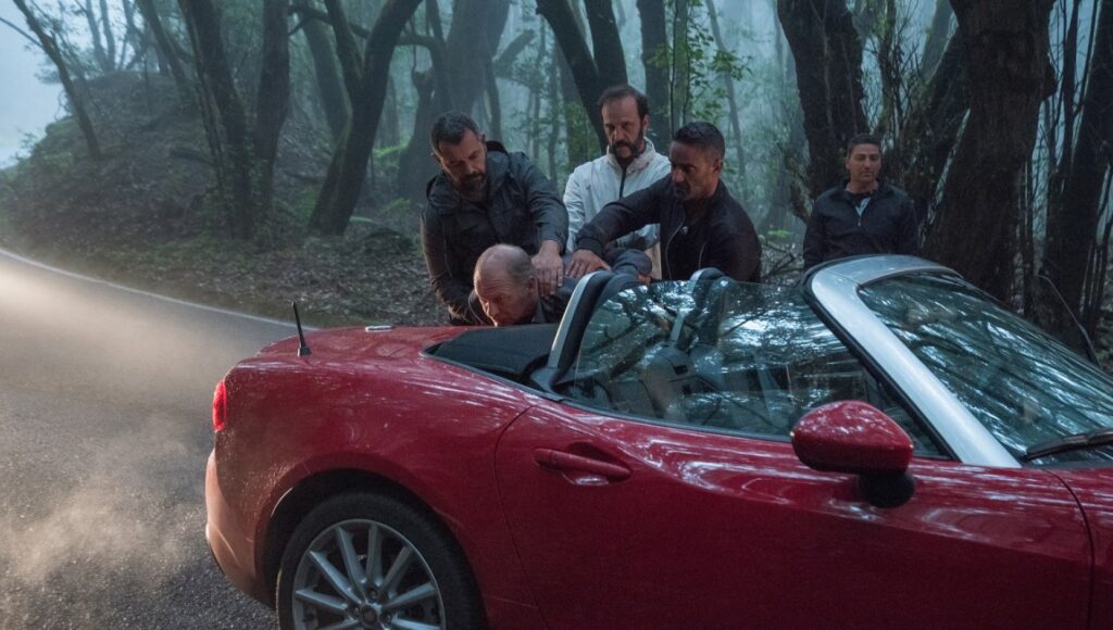 The Whistlers movie scene: Men surrounding a red convertible car in a wooded area.