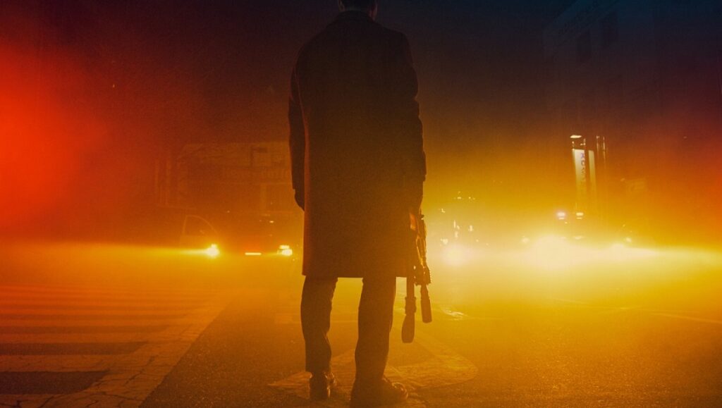 Time to Hunt | Yoon Sung-hyun Time to Hunt movie scene. Man in coat stands on a crosswalk in a hazy, orange-lit street.