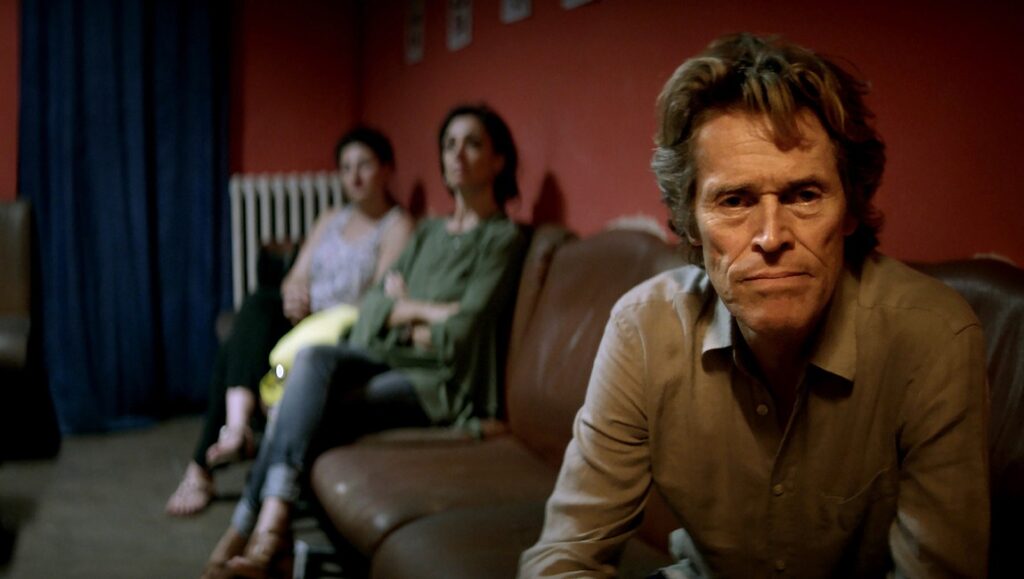 Willem Dafoe in Tommaso by Abel Ferrara. Dafoe sits in the foreground, two women on a couch behind him.