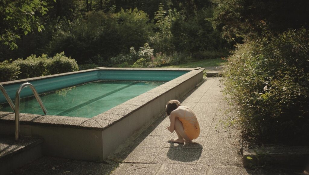 The Trouble with Being Born film still: Child squats by a pool in a lush, green outdoor setting.