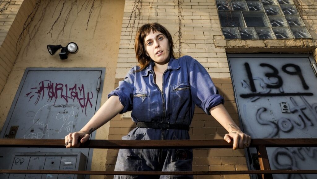 U.S. Girls Heavy Light album art. Musician in blue jumpsuit on balcony with graffiti-covered doors.