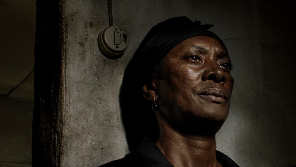 Vitalina Varela, Pedro Costa film. Portrait of Vitalina, head wrapped, looking up in a dark, textured interior.