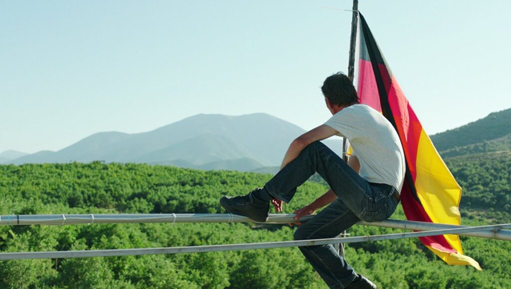 Western film still. Man sits on a fence with a German flag, overlooking a green landscape.