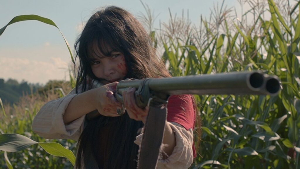 Birdshot movie scene: A girl fiercely aims a shotgun in a cornfield, hinting at the film's dark themes.