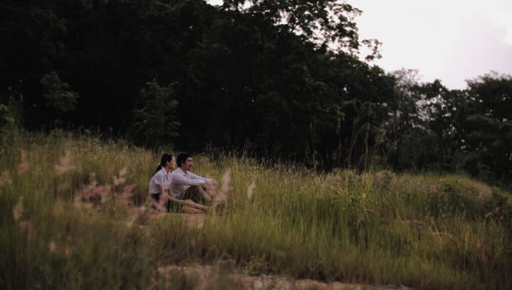 By the Time It Gets Dark film still. Two people sit in a field of tall grass, with trees in the background. Anocha Suwichakornpong film.