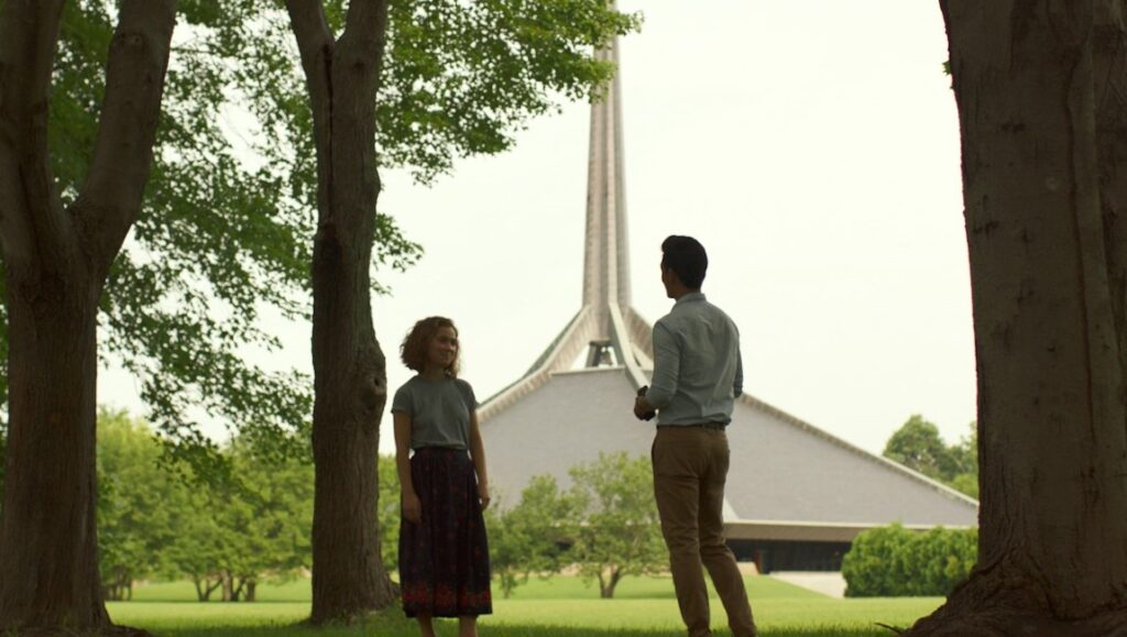 Scene from Columbus film: A man and woman stand in a park with a modern building in the background.