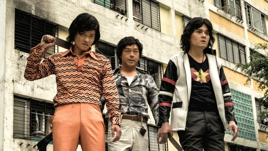 Dealer/Healer | Lawrence Lau Dealer/Healer film still. Three Asian men in 70s attire stand before a building.