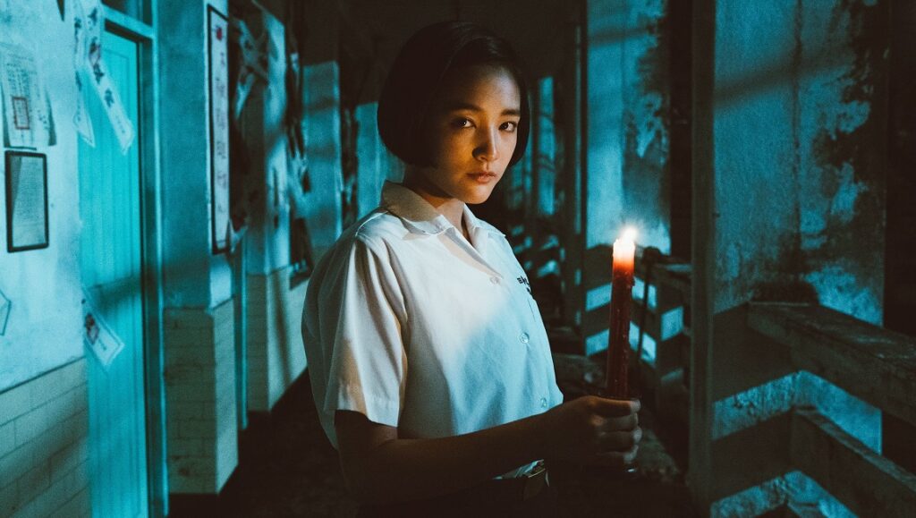 Detention | John Hsu Detention movie scene: A student holds a candle in a dark school hallway. Horror film vibes.