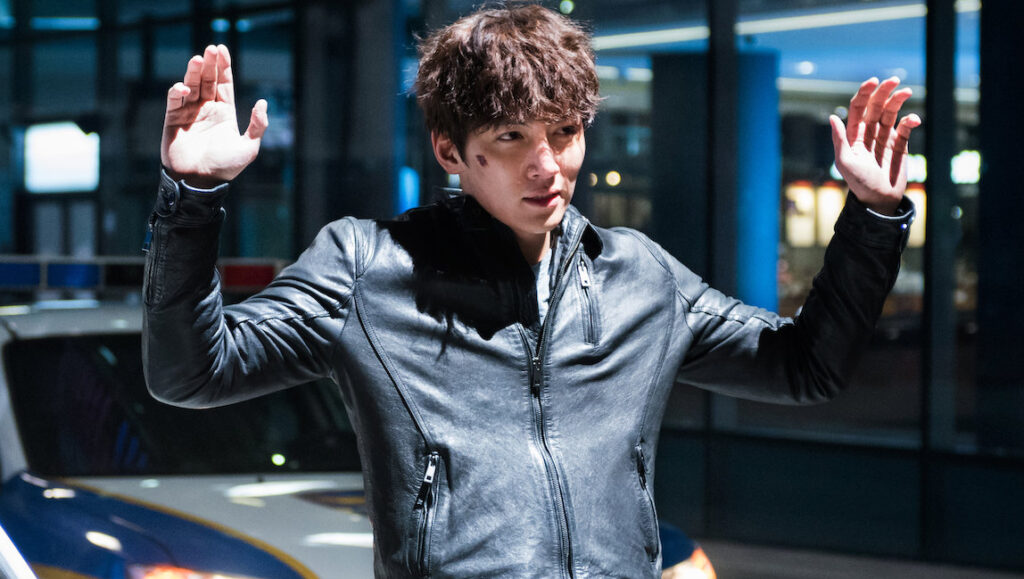 Fabricated City movie scene: Man in leather jacket with hands up, police car. Park Kwang-hyun film.