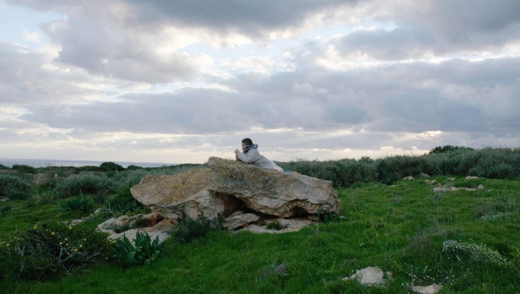 Fire at Sea still: Boy rests on rock, green field, cloudy sky. Gianfranco Rosi film scenery.