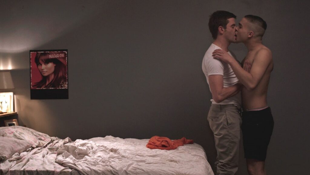 Four Days in France film still. Two men kiss intimately in a bedroom with a messy bed.