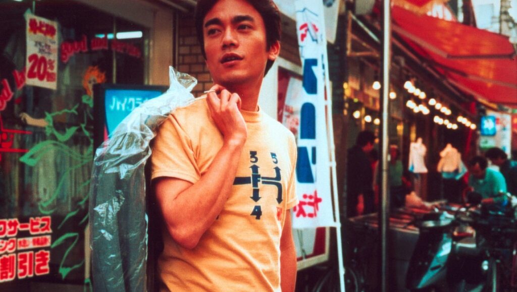 Hush! — Ryosuke Hashiguchi Ryosuke Hashiguchi's Hush! film still. Man in yellow shirt stands on a busy street, looking thoughtful.