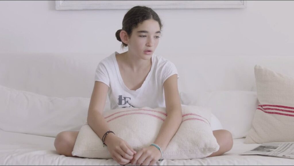 Still from Kékszakállú by Gastón Solnicki: Young woman sits cross-legged on a white sofa, looking contemplative.