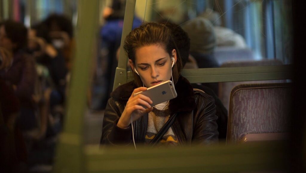 Kristen Stewart in Personal Shopper movie. Woman with headphones using phone on public transport.