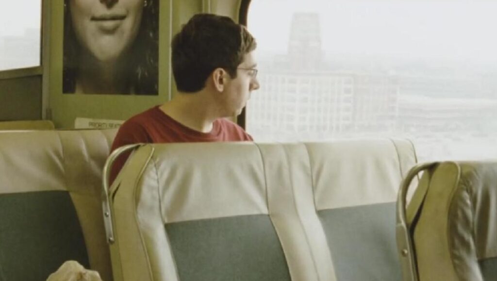 Short Stay | Ted Fendt Short Stay film still: Man on train looks out window at city. Poster on wall.
