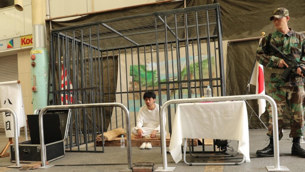 Artist of Fasting film still. Masao Adachi movie scene with a caged man, soldier, and Japanese flag.