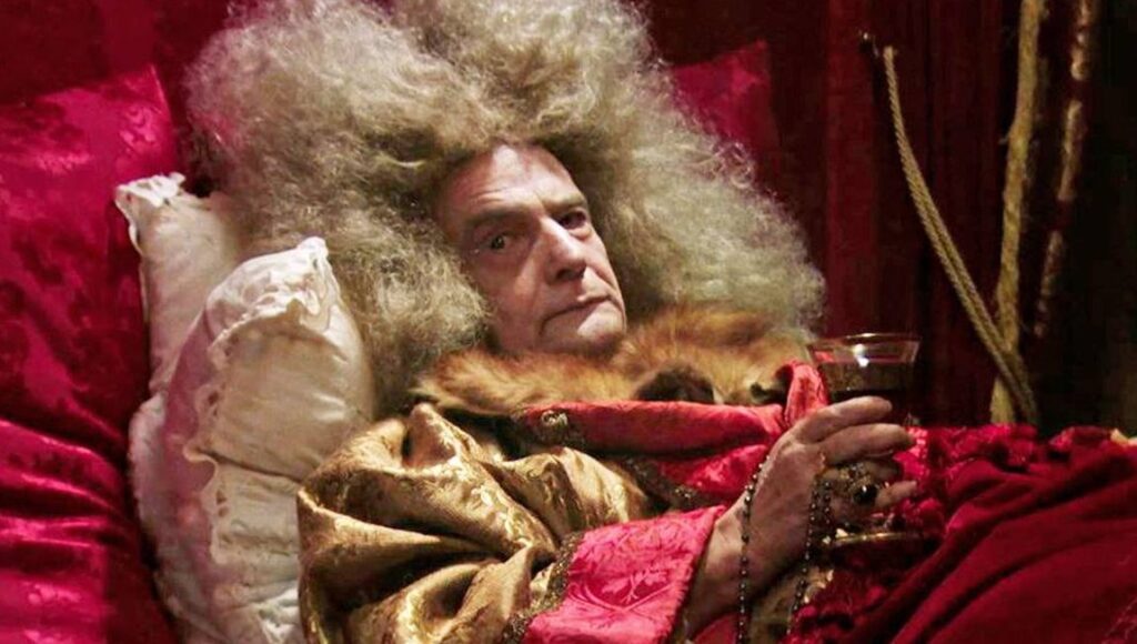 Still from The Death of Louis XIV film. Man with wig, holding wine, lying in bed. Albert Serra movie.