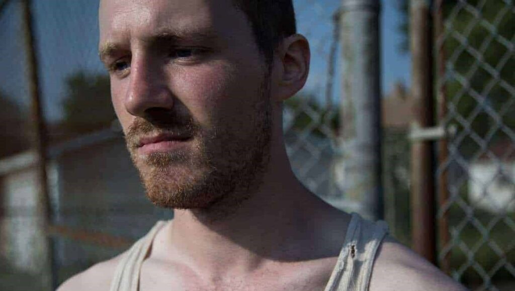 Ashley McKenzie's Werewolf: man with a beard in a tank top near a chain link fence.