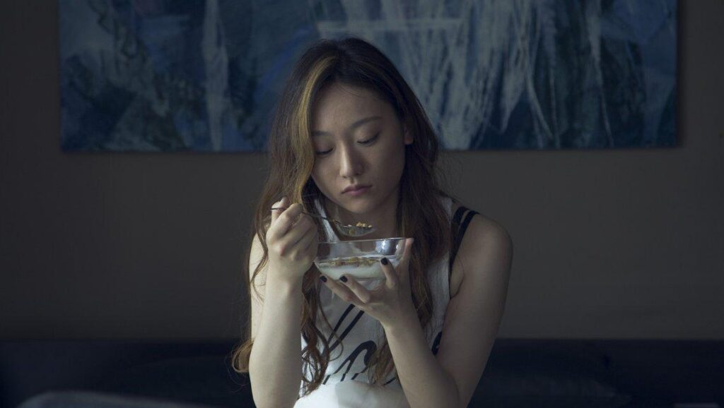 Takuro Nakamura's West North West: Woman eating cereal. Indoor scene with muted tones. Art on wall.