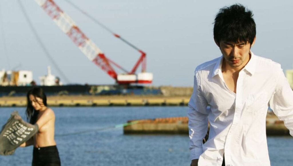 Wet Woman in the Wind film still. Akihiko Shiota film features a woman and man near water with a crane in the background.