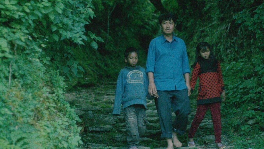 White Sun film still. Man holding hands with a boy and girl walking on a stone path surrounded by lush greenery.