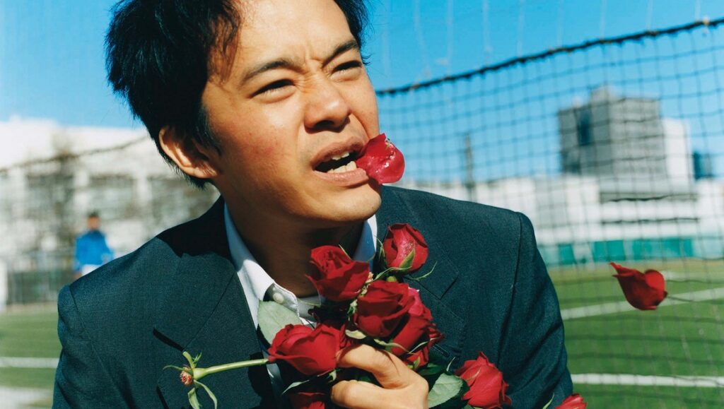 Tetsuya Mariko's Miyamoto still. Man in suit with red roses, one in mouth. Quirky, artistic film shot.