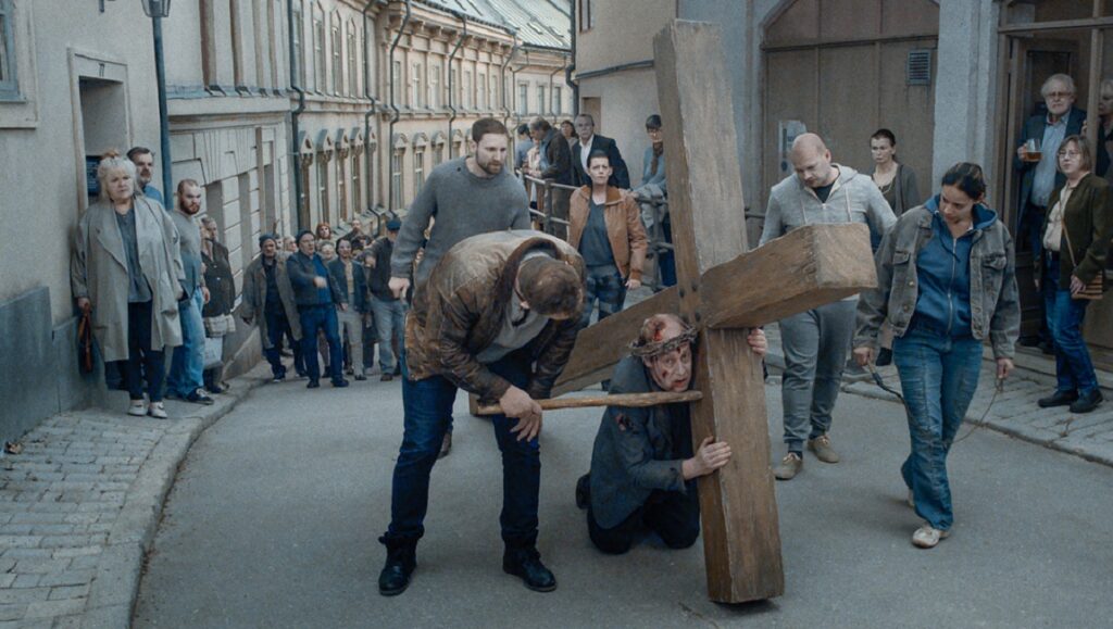 About Endlessness film still: Man carrying a large cross with help, crowd watching. Roy Andersson film.