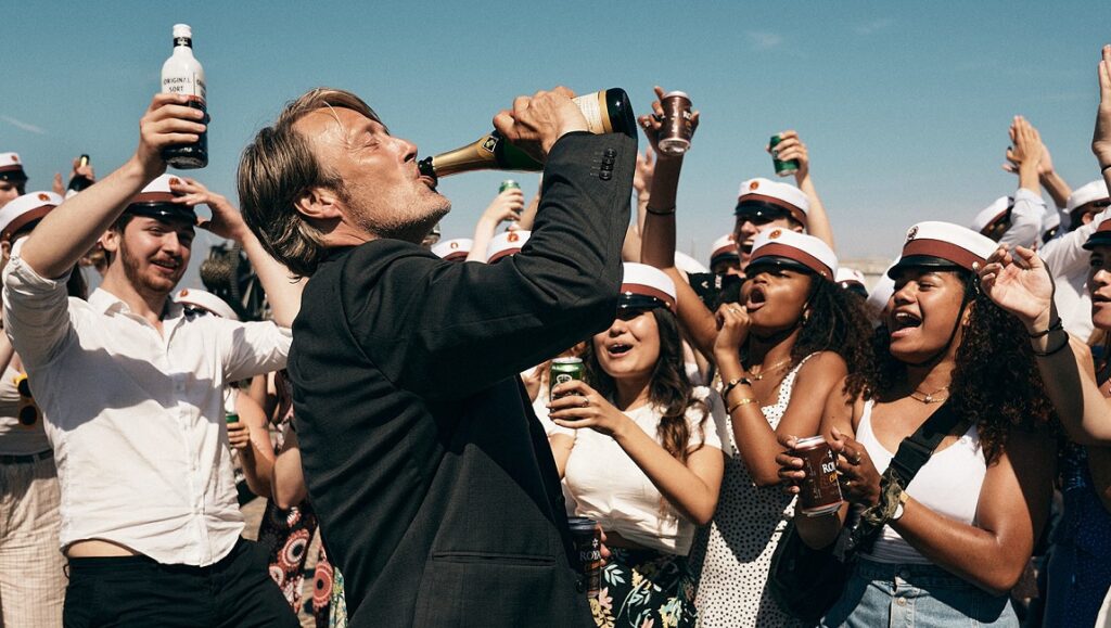 Another Round | Thomas Vinterberg Mads Mikkelsen drinks in Another Round movie scene. Crowd celebrates with drinks, wearing graduation caps. Thomas Vinterberg film.