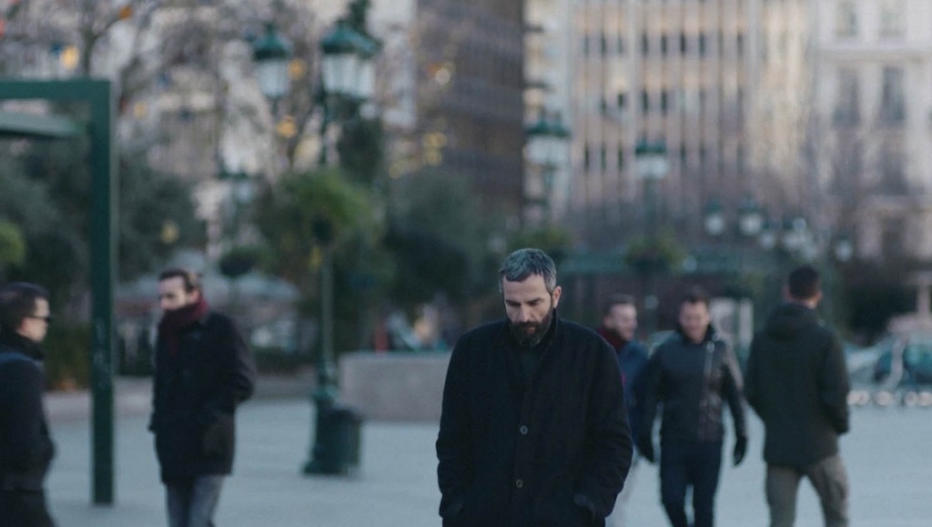 Apples — Christos Nikou Christos Nikou's Apples: Man in black coat walks in city square, surrounded by blurred figures. Urban, melancholic scene.