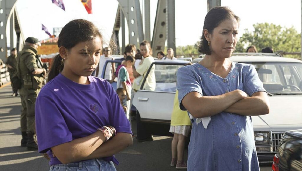 Beans movie still from TIFF 2020. Two Indigenous women stand with arms crossed. Cars and soldiers in the background.