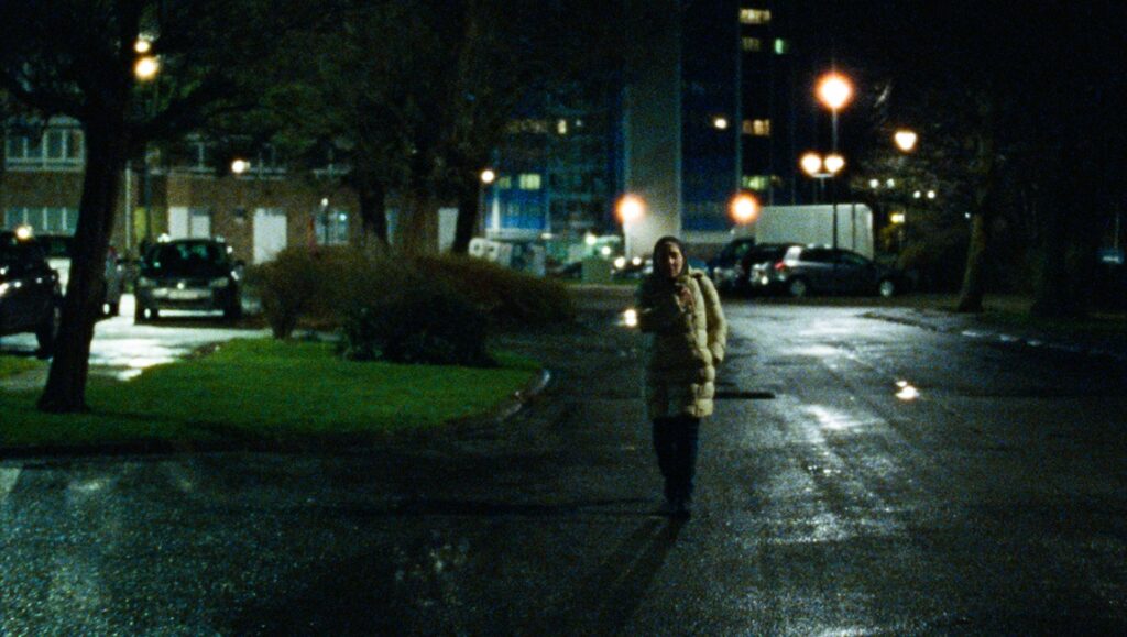 Ghost Tropic | Bas Devos Still from Before We Vanish (September 2020): Woman walks on a dark, wet street at night. Streetlights glow.