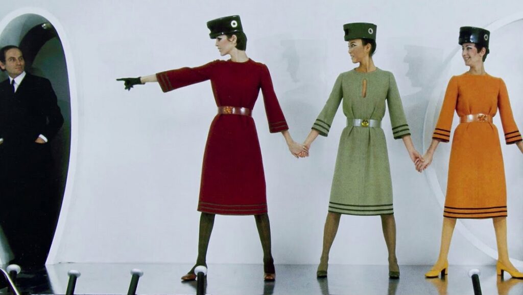 House of Cardin film still. Models in 1960s-style dresses and hats hold hands. A man in a suit watches.