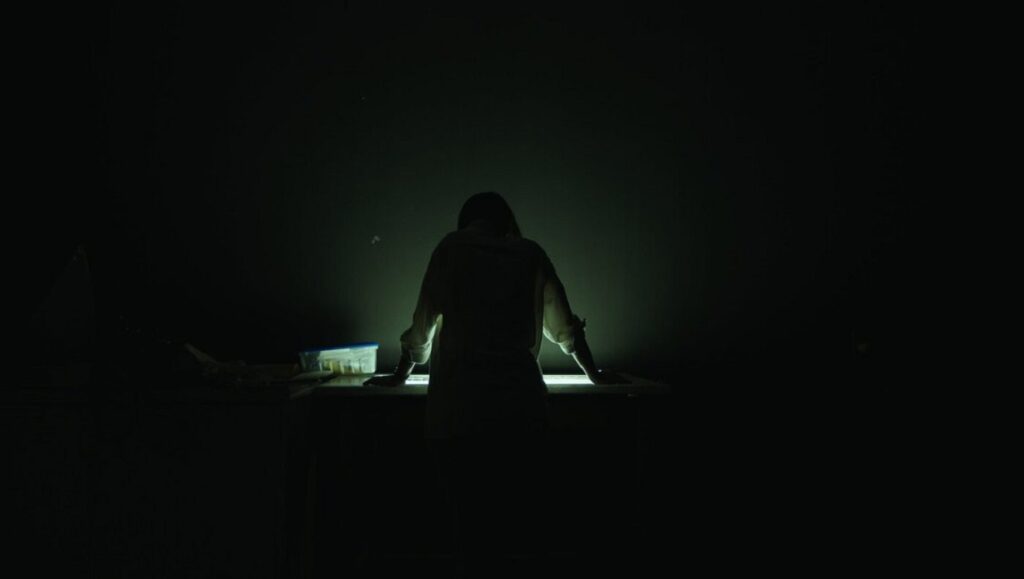Still Processing by Sophy Romvari: Silhouette of a person at a desk in the dark, illuminated by a light source.