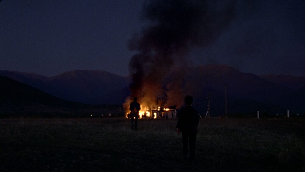 Still from Red, White and Blue NYFF film. Two figures watch a burning building at night, mountains in the background.