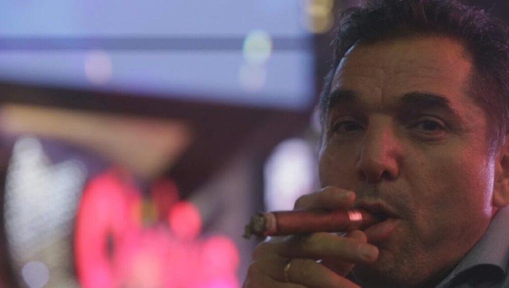 The Projectionist | Abel Ferrara Abel Ferrara in The Projectionist movie. Man smoking a cigar, with blurred lights in the background.