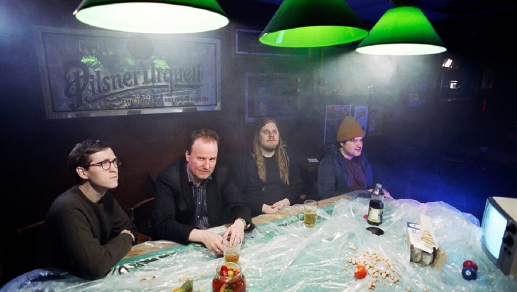 Ledger Line Q3 2020: Protomartyr band members at a table with drinks and snacks in a dimly lit bar setting.