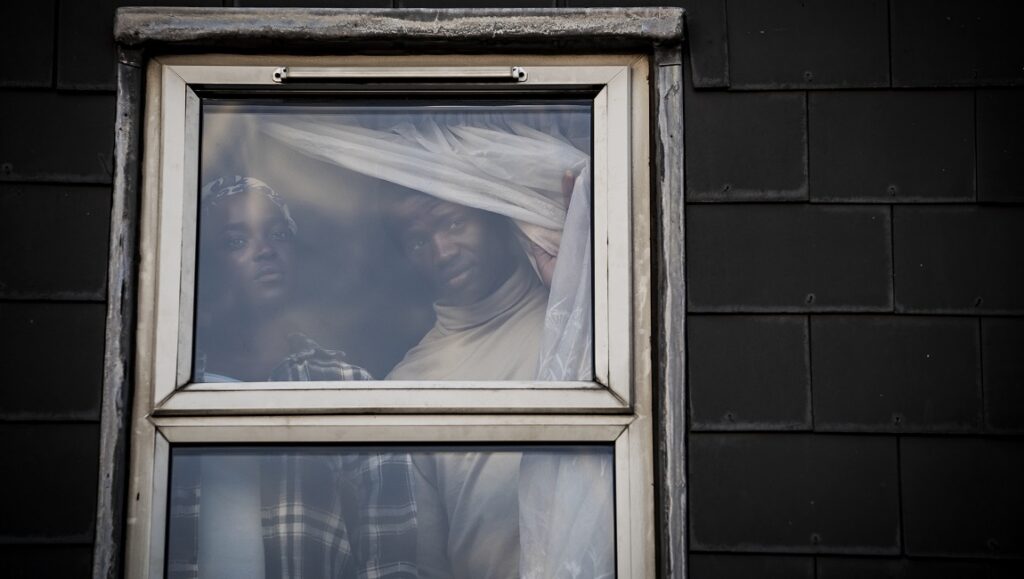 His House | Remi Weekes His House movie still. A man and woman peer out of a window, faces partially obscured by a sheet.