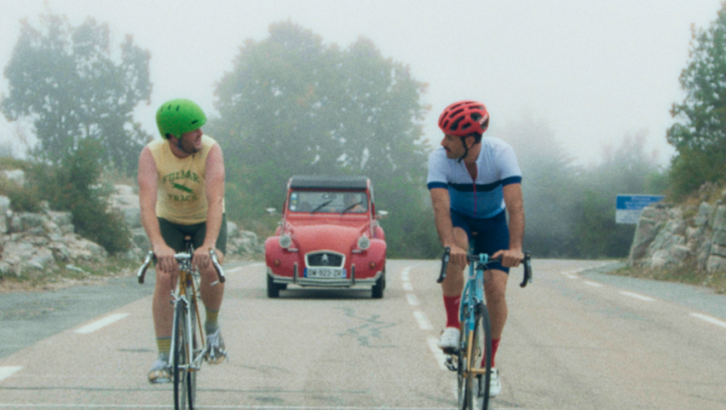 The Climb movie scene. Two cyclists ride uphill with a vintage red car behind them.