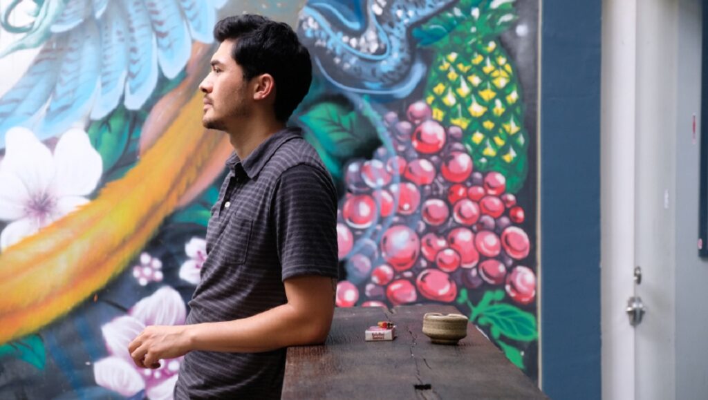 Hong Khaou's Monsoon: Man with mural art backdrop. Pineapple, grapes, and flowers adorn the wall. Film scene.