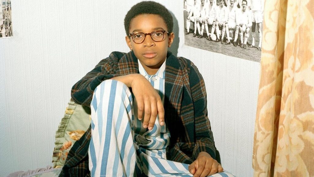 Still from Education by Steve McQueen. A young boy in glasses, pajamas, and robe sits on a bed.