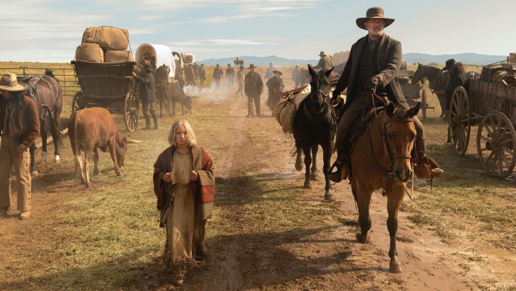 News of the World | Paul Greengrass News of the World movie scene. Tom Hanks rides a horse alongside a girl in a Western landscape with wagons.