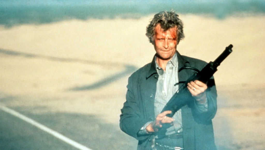 Richard Harmon as The Hitcher holding a shotgun. Blood on face, standing on a desert road.