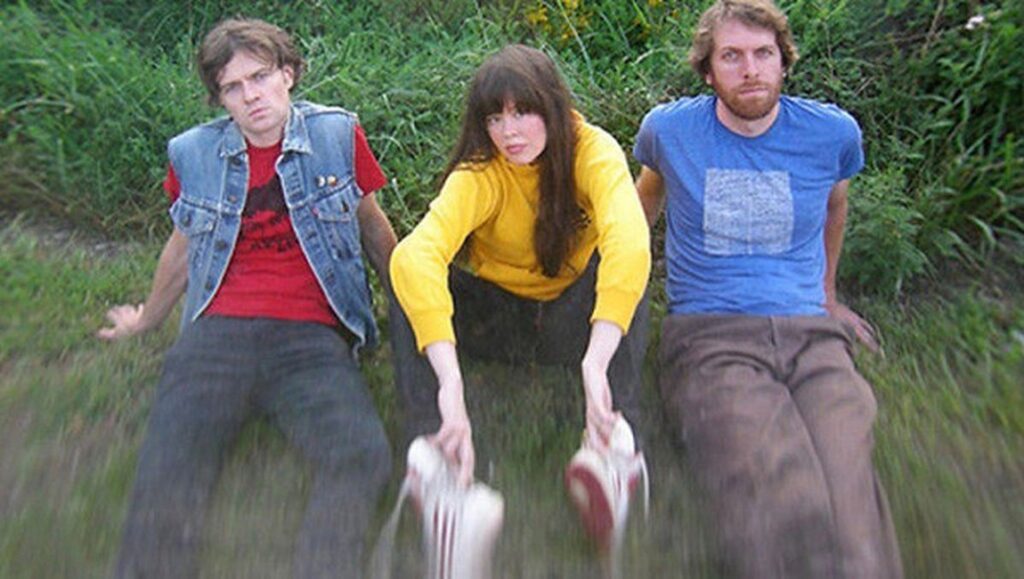 Magic Markers band, 2020. Three members sit in grass, holding shoes. Indie rock music group.