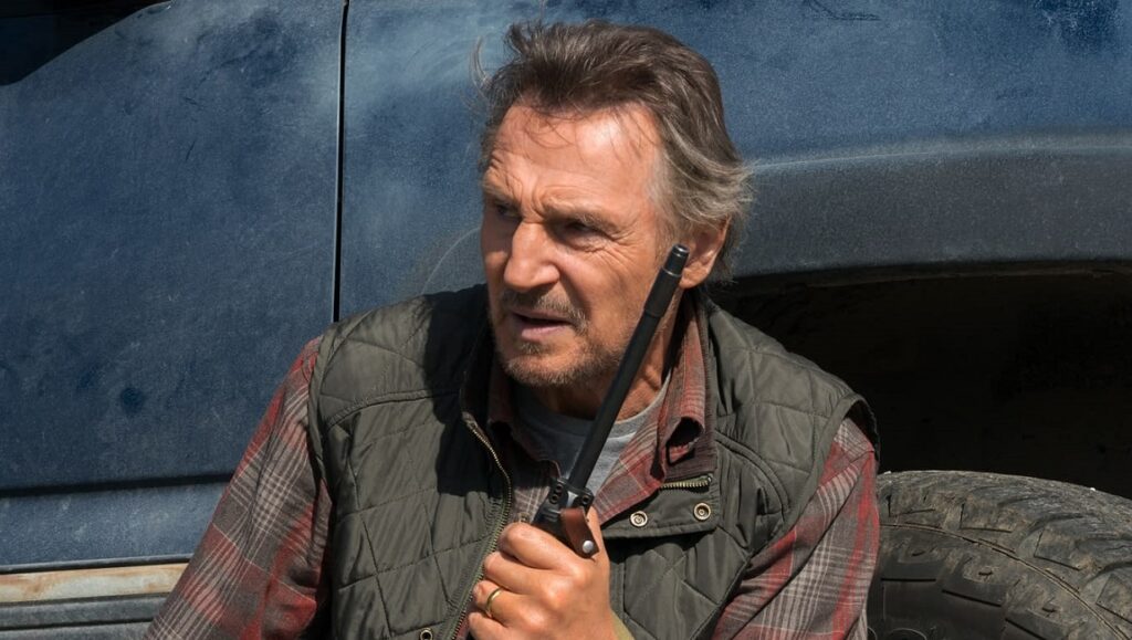 Liam Neeson in The Marksman. Actor holding a radio, wearing a vest and plaid shirt. Movie still.